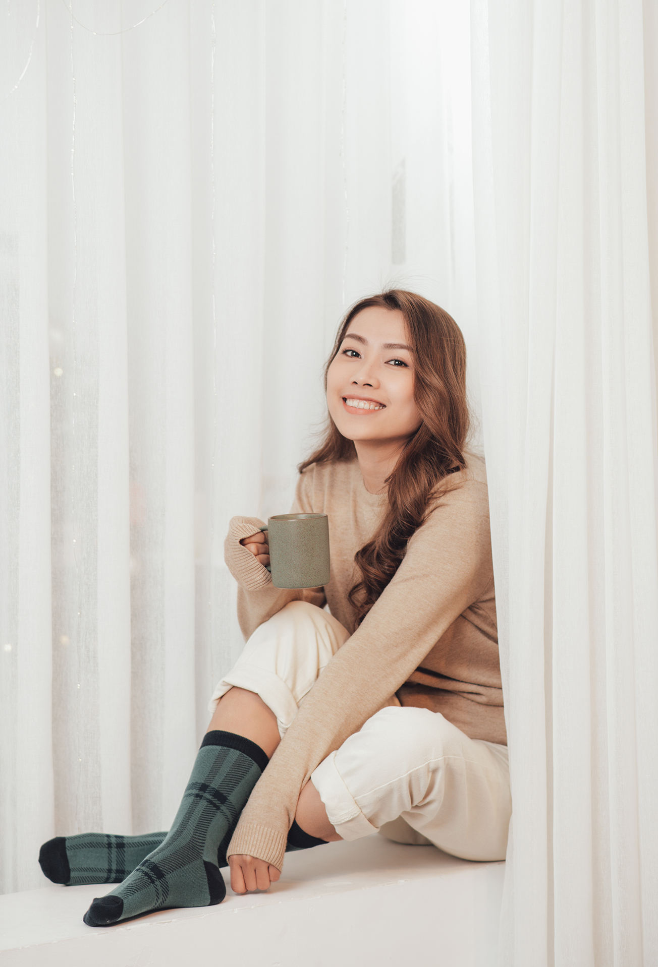 Happy beautiful woman drinking hot coffee sitting on window sill in christmas decorated home. Holiday concept